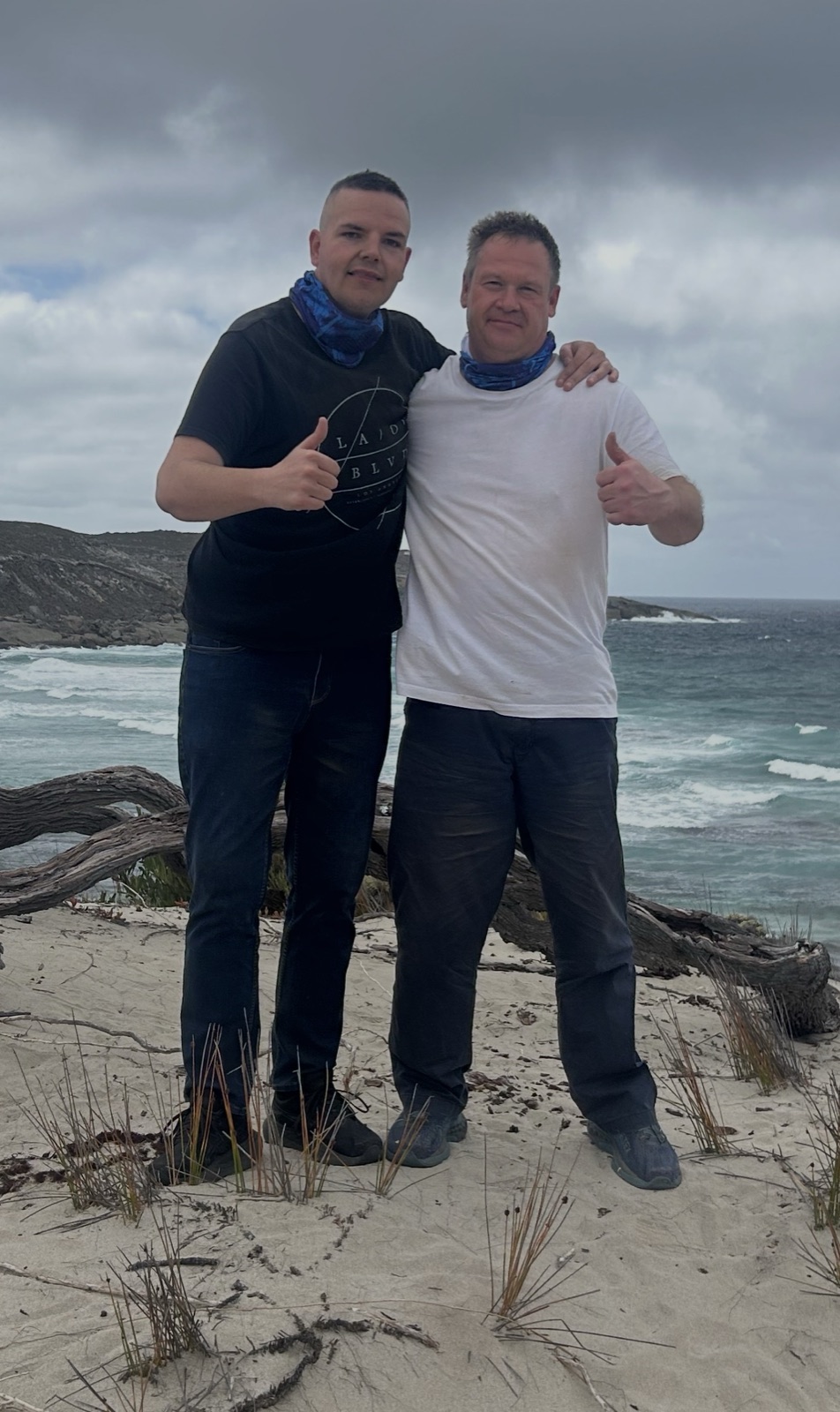 Two of the Equilibrium community arm in arm on the beach, both giving a thumbs up under a dramatic sky
